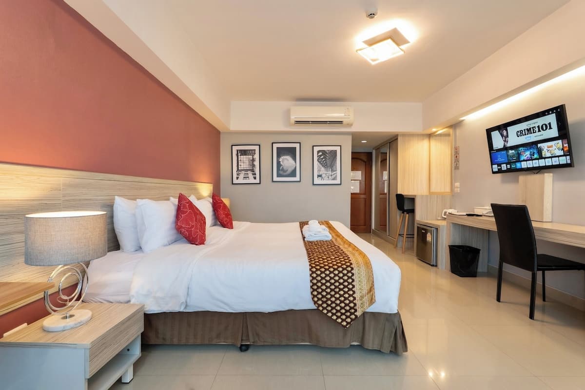 A hotel room with a bed, bedside table, lamp, TV, and a desk with a chair.