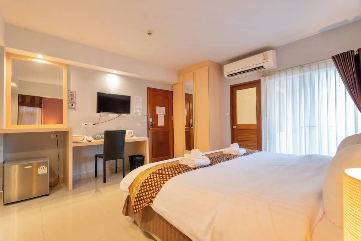 A hotel room with a bed, desk, chair, TV, air conditioning unit, and a small trash bin.