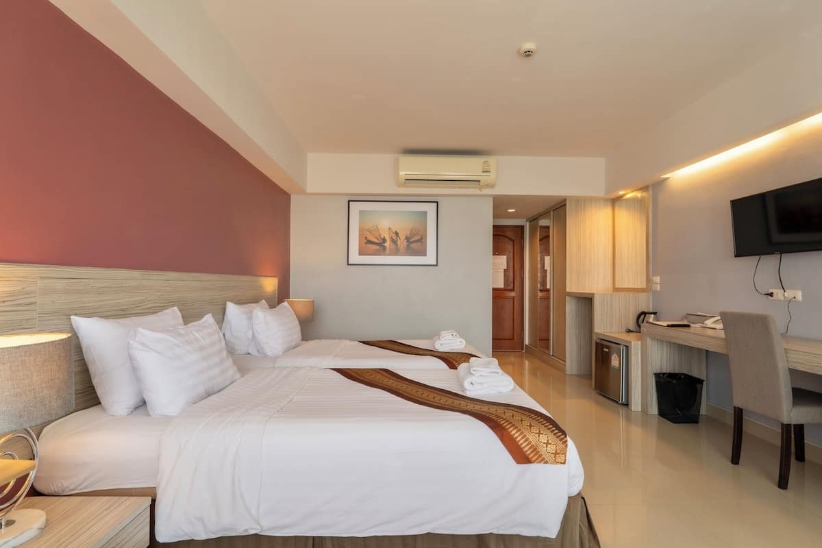 A hotel room with a large bed, a desk, a chair, a TV, and an air conditioning unit.