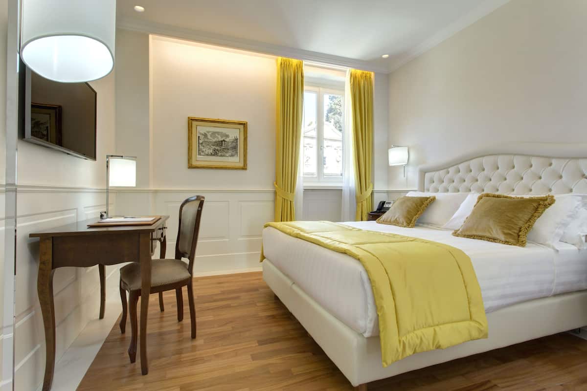 A bedroom with a large bed, a desk, and a chair. There is a window with curtains, a framed picture on the wall, and a lamp on the desk.