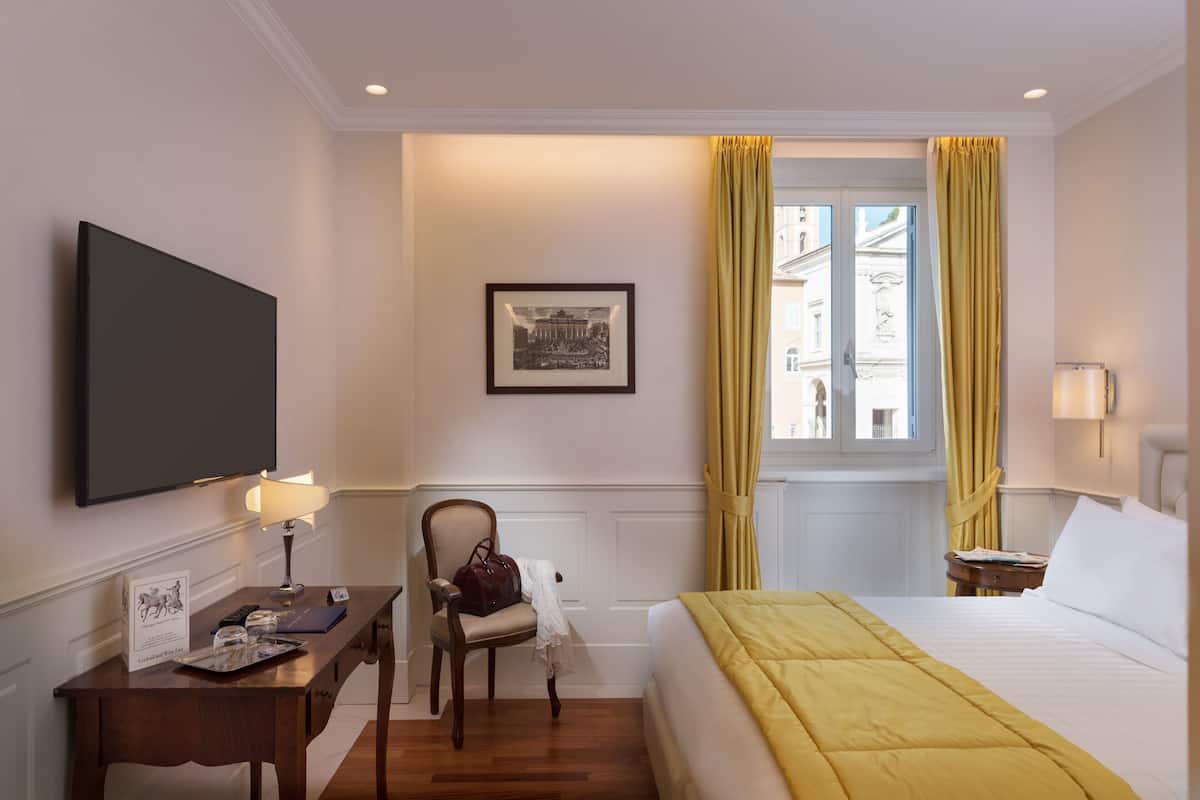 A hotel room with a bed, a desk with a lamp, a chair, a framed picture on the wall, and a window with curtains.