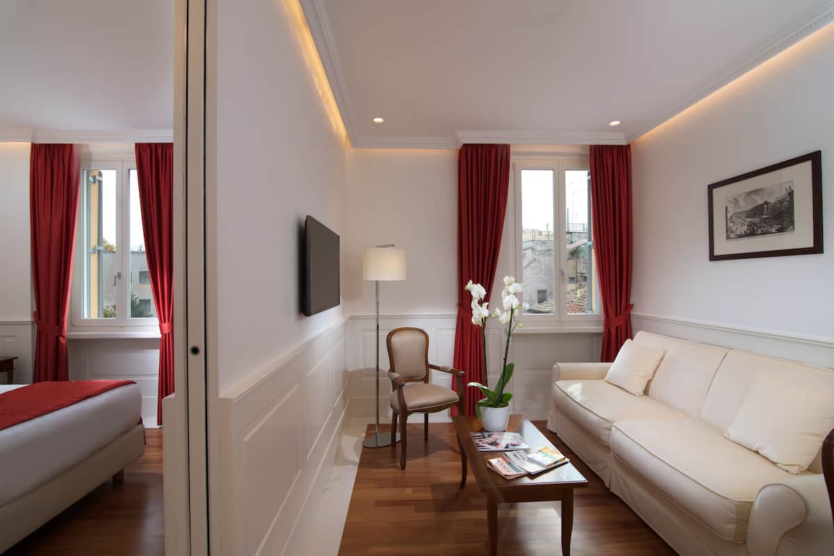 A hotel room with a bed, a sofa, a coffee table, a chair, a TV, and a window with curtains.