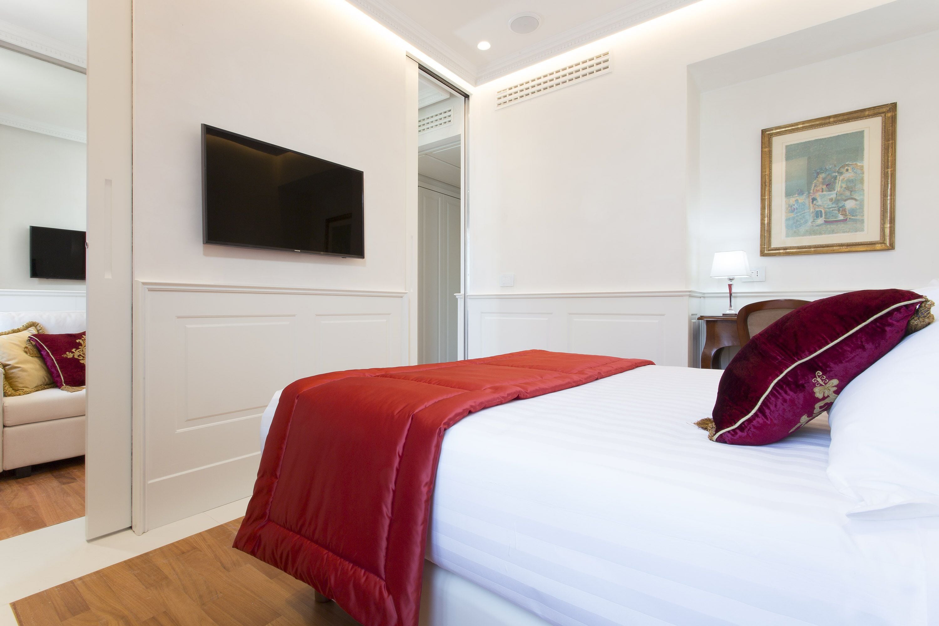 Une chambre d’hôtel avec un lit, un téléviseur fixé au mur, un tableau et une porte donnant sur une autre pièce.
