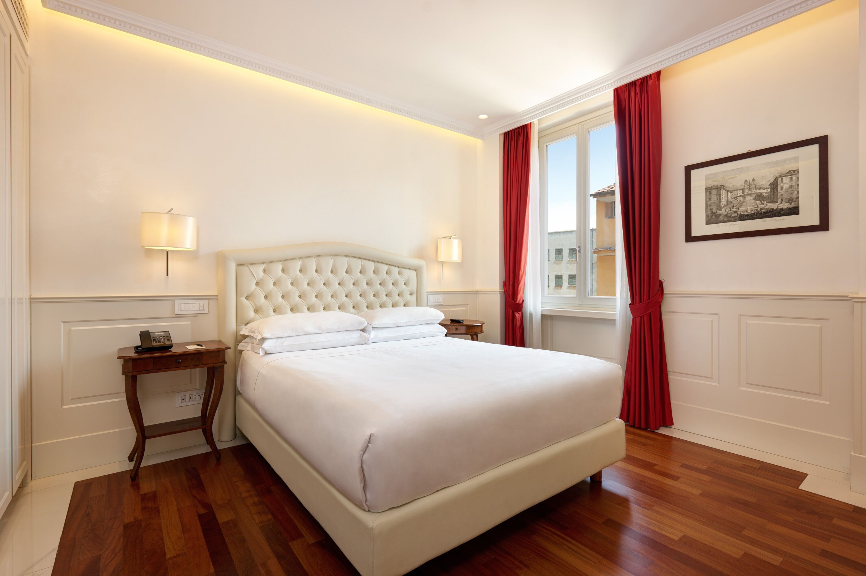 A hotel room with a large bed, two bedside tables, a nightstand, a window with red curtains, and a framed picture on the wall.