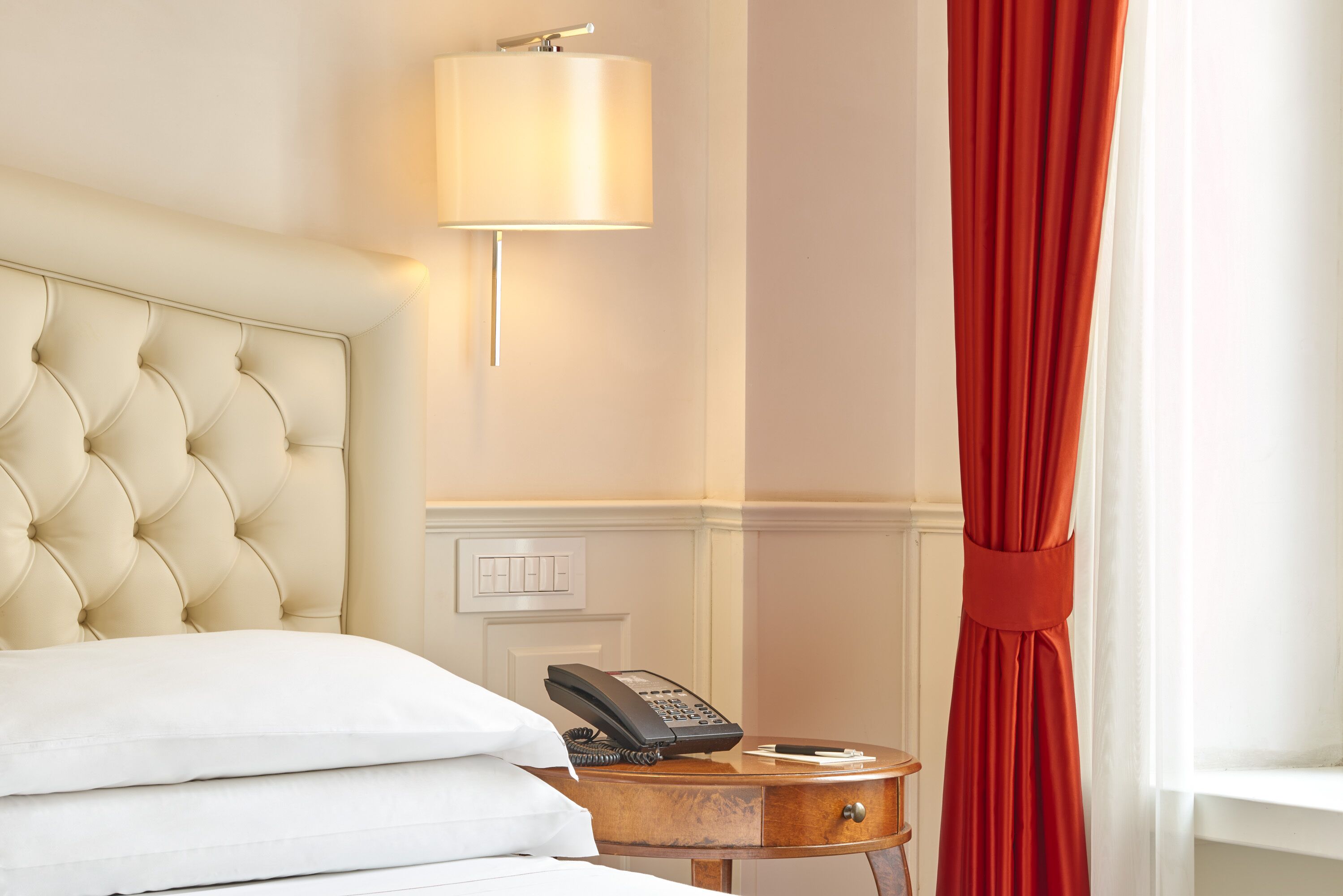 Quarto de hotel com cabeceira estofada, telefone de mesa, uma pequena mesinha de cabeceira e cortina vermelha.