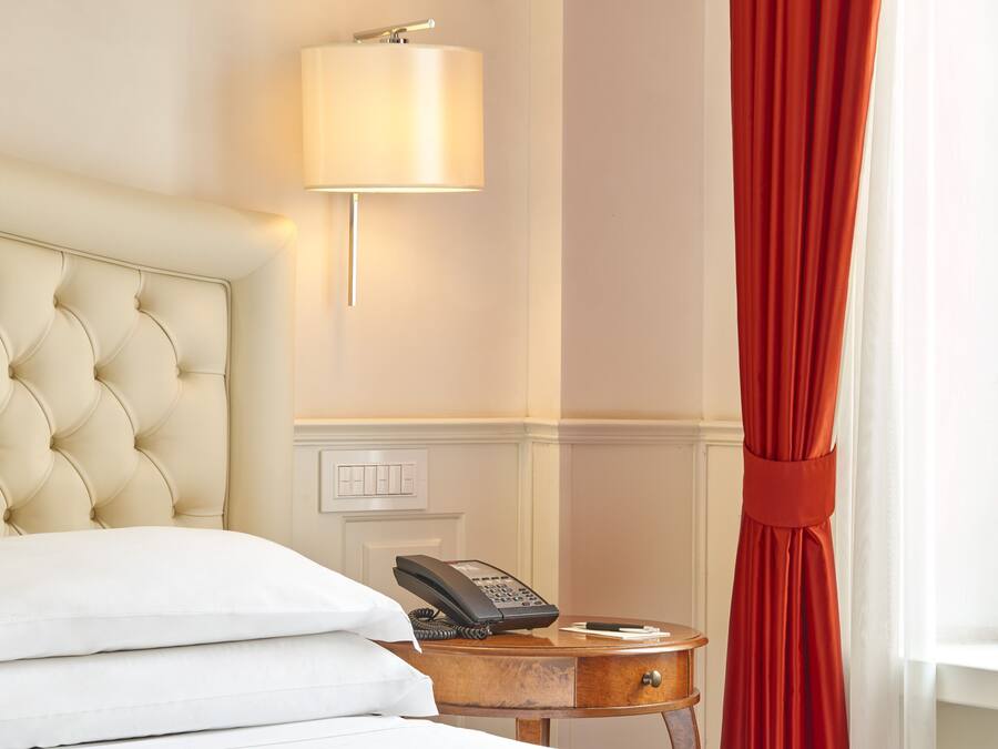 Quarto de hotel com cabeceira estofada, telefone de mesa, uma pequena mesinha de cabeceira e cortina vermelha.