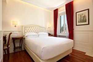 Una camera d'albergo con un letto grande, due lampade ai comodini, una sedia di legno, un tavolino e un quadro appeso alla parete.