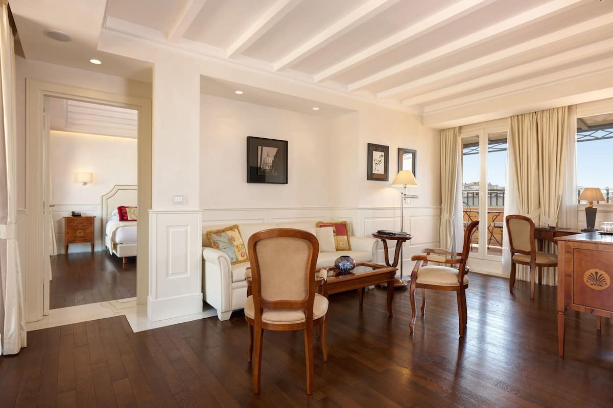 A hotel room with a wooden floor, a white sofa, a wooden chair, a small table, and a desk with a lamp.