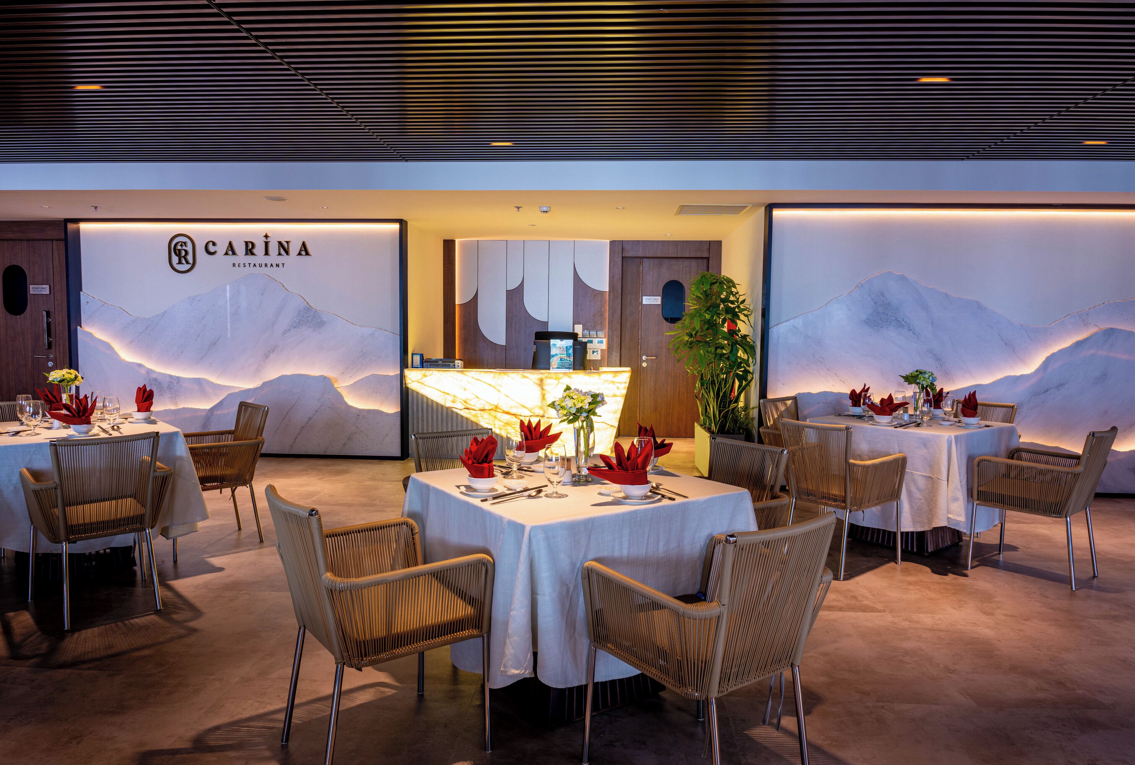Un restaurante con mesas redondas y sillas de mimbre, un muro con motivos de montaña y una zona de recepción.
