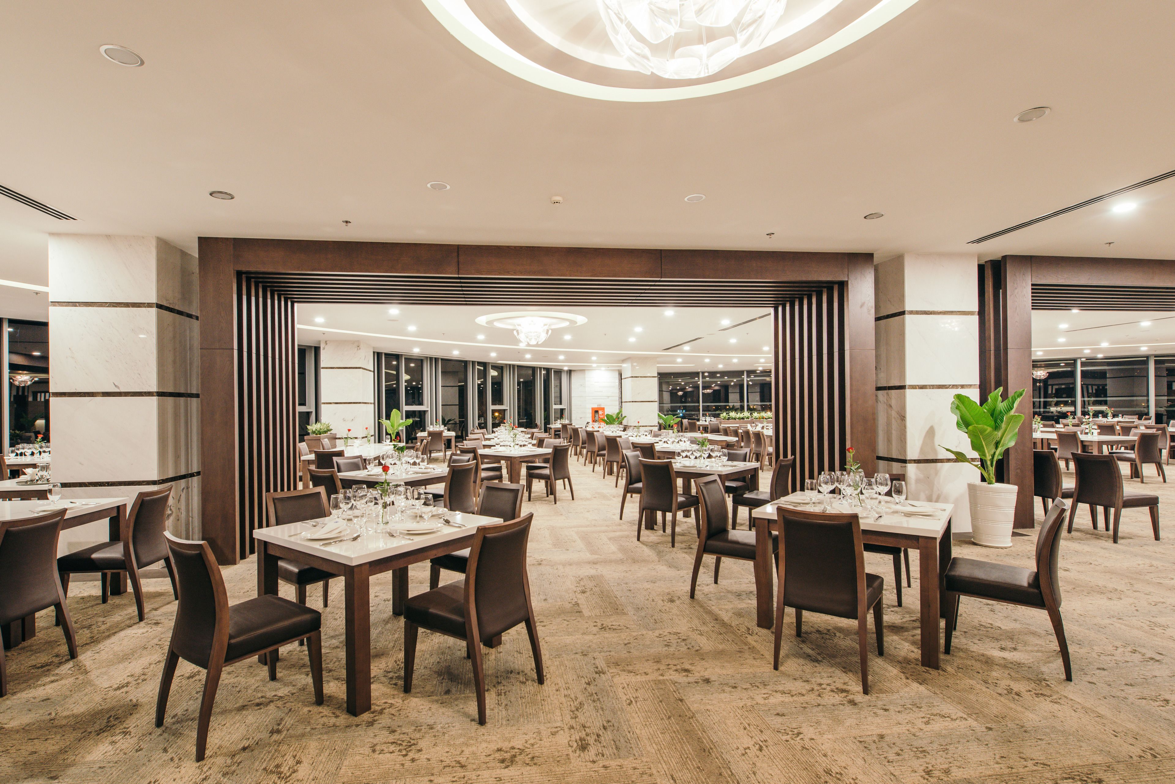 A spacious dining area with multiple tables and chairs, large windows, and a modern ceiling design.