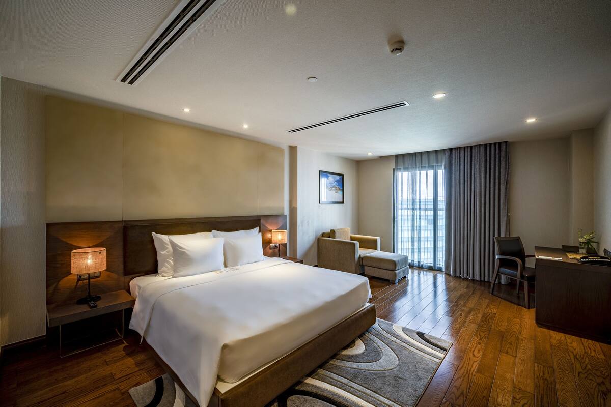 Ein Hotelzimmer mit einem großen Bett, einem Schreibtisch, einem Sessel, einer Couch und Blick ins Freie.