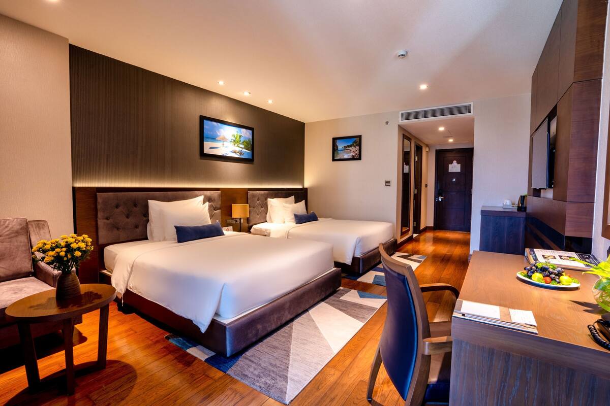 Ein Hotelzimmer mit zwei Betten, einem Schreibtisch, einem Sessel, einem kleinen Tisch mit Blumen und einem Esstisch mit Obstteller.