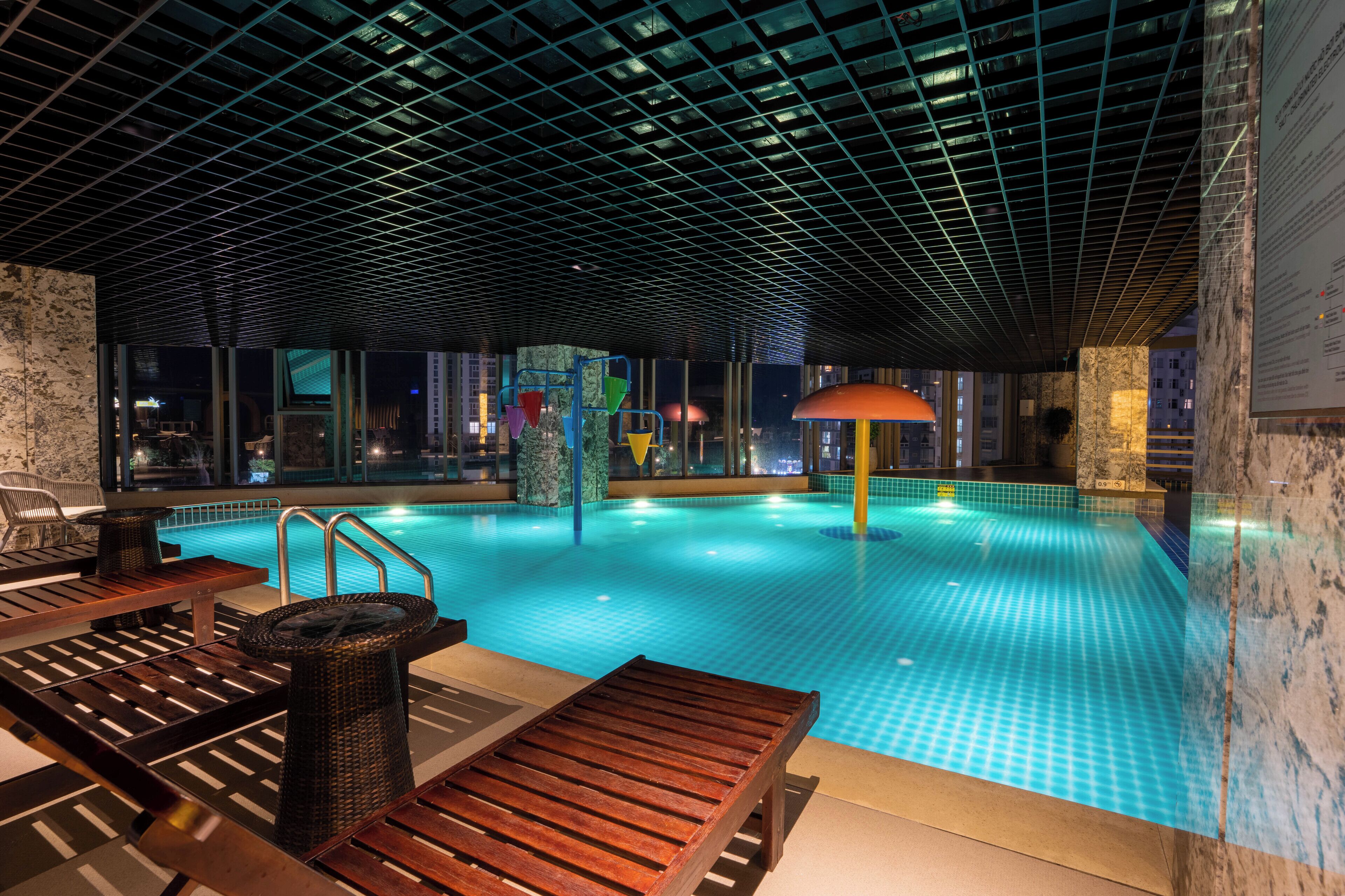 Piscina interna com piso ladrilhado, espreguiçadeiras de madeira e teto com padrão de grade.
