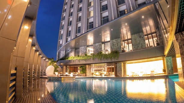 A modern hotel with a pool, a curved glass facade, and a well-lit lobby area. -  The Malibu Hotel (Ho Chi Minh City)