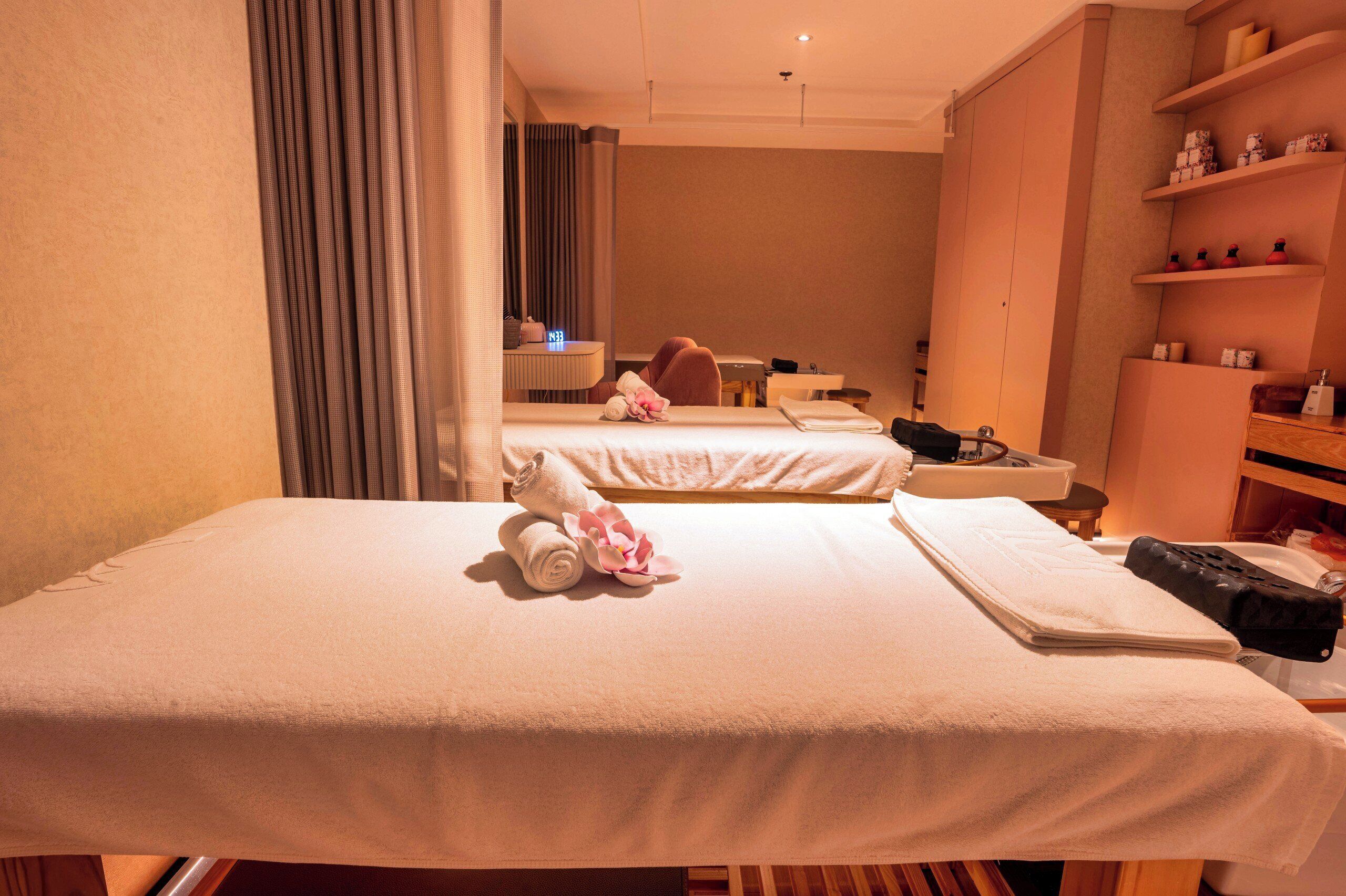 A hotel room with a bed, a massage table, a small desk with a lamp, and a shelf with toiletries.