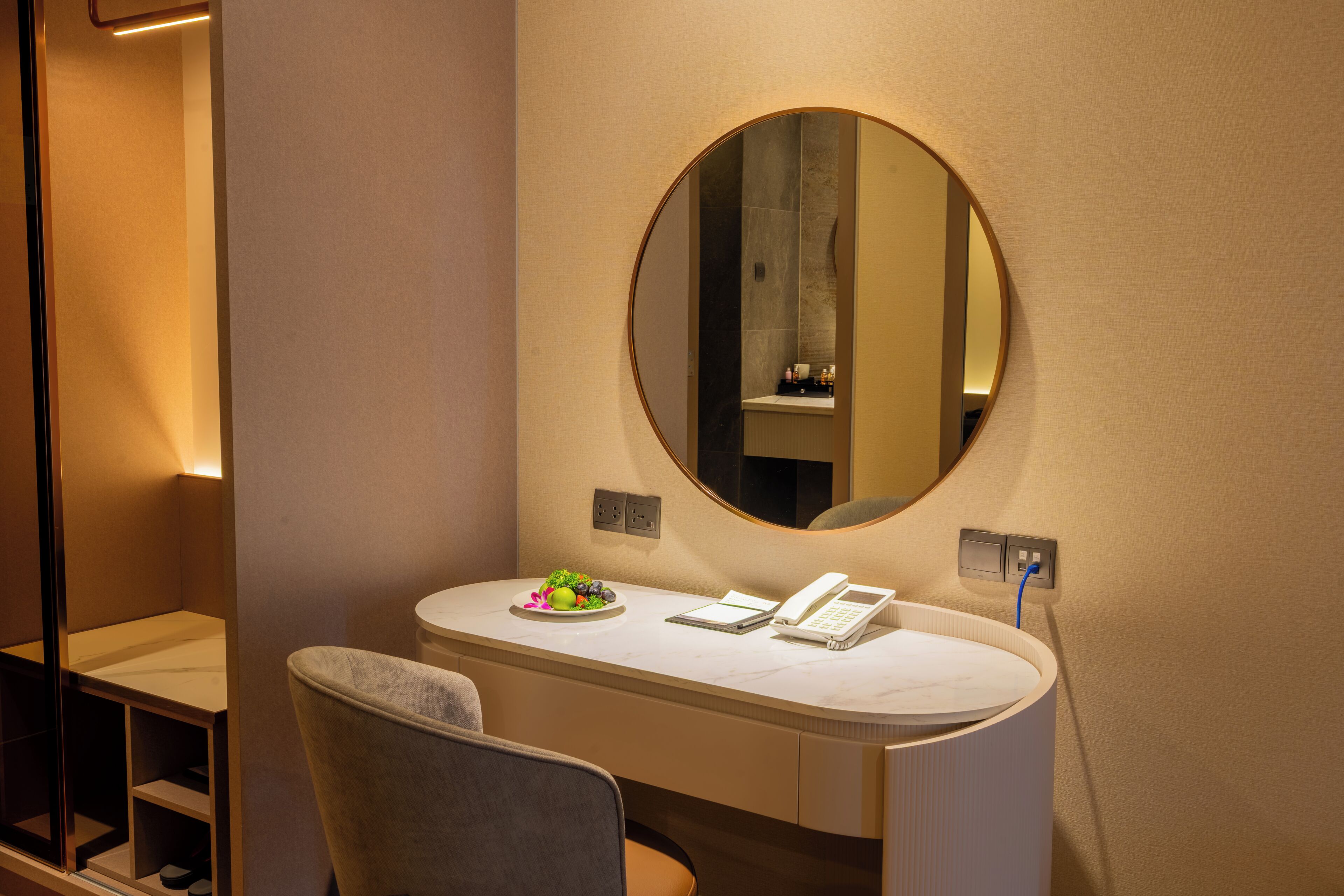 A hotel room with a vanity, a mirror, a chair, and a small table with toiletries.
