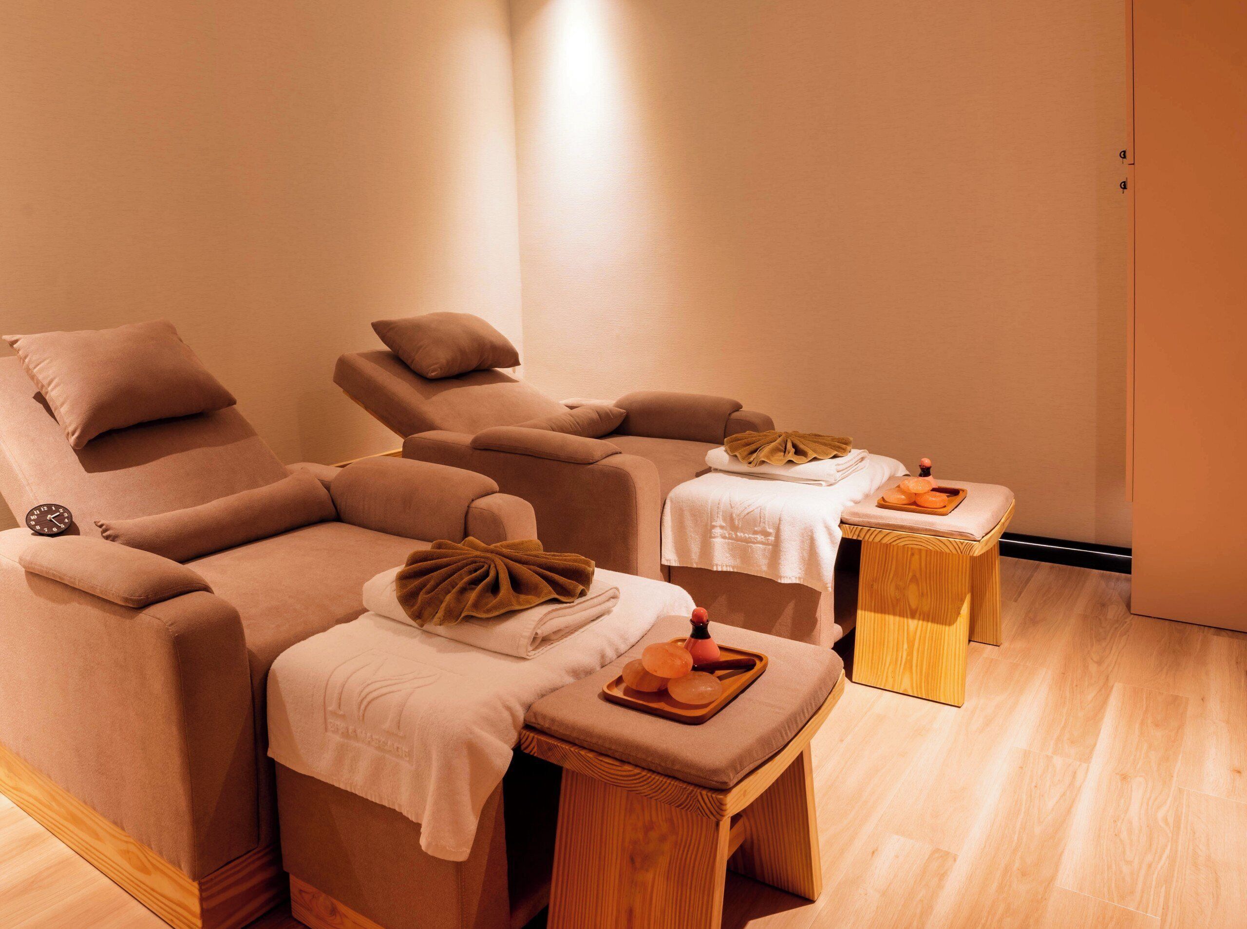 A massage room with two reclining chairs, a small wooden table with a towel and a tray of oranges, and a clock on the wall.
