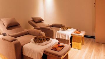 A massage room with two reclining chairs, a small wooden table with a towel and a tray of oranges, and a clock on the wall.