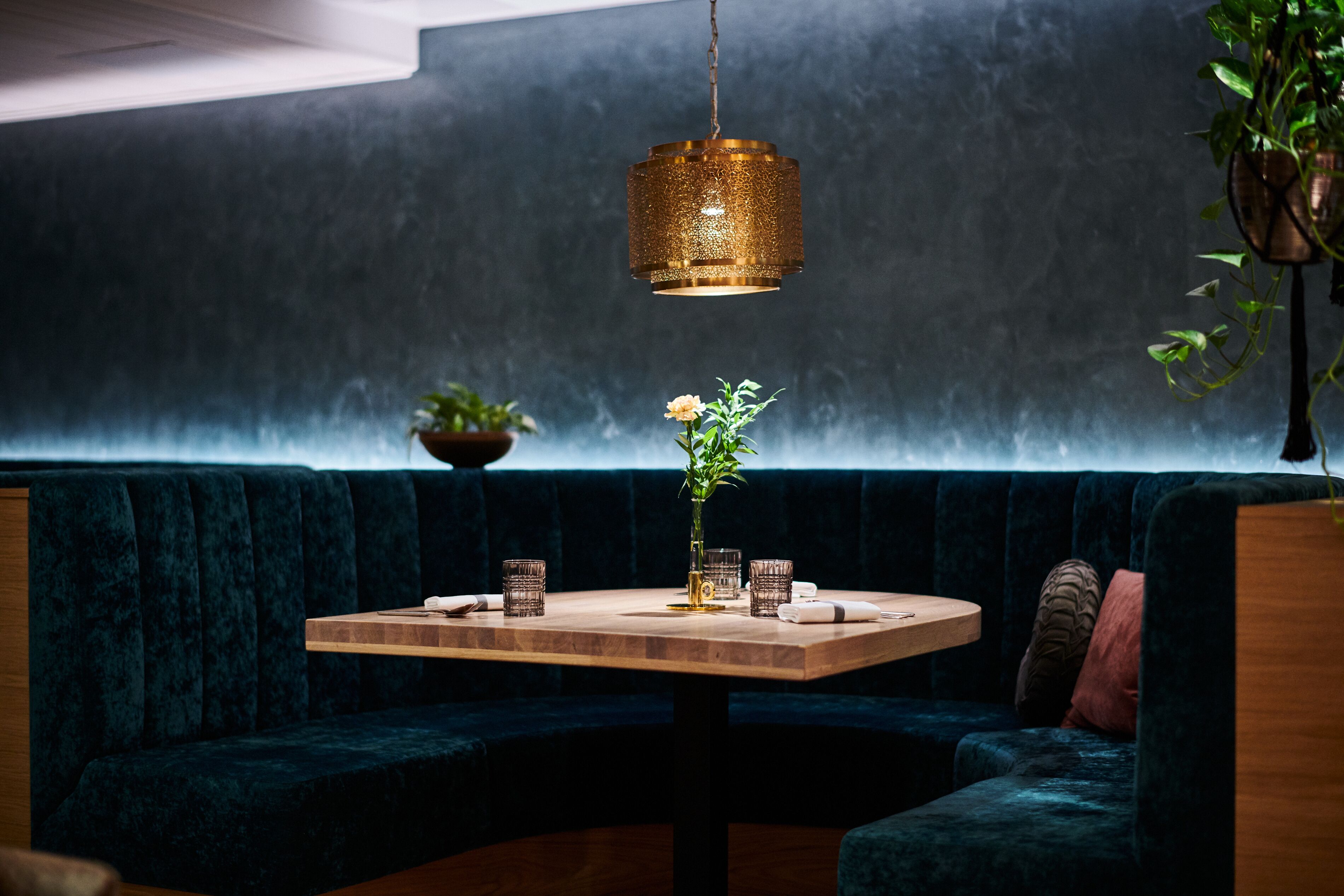 A cozy seating area with a dark blue velvet sofa, a wooden table with glassware, and a hanging gold pendant light.