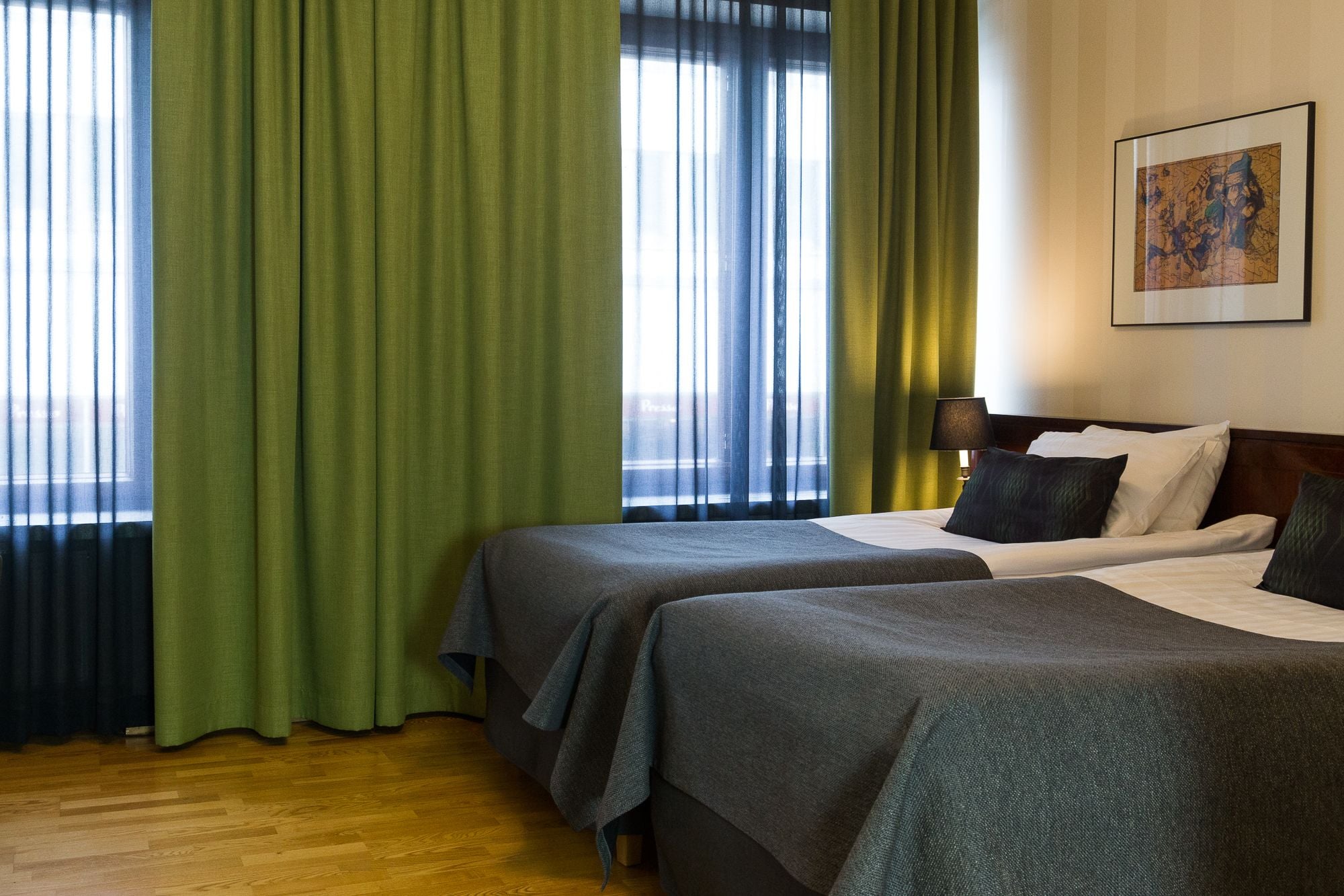A hotel room with two beds, a wooden floor, green curtains, and a painting on the wall.