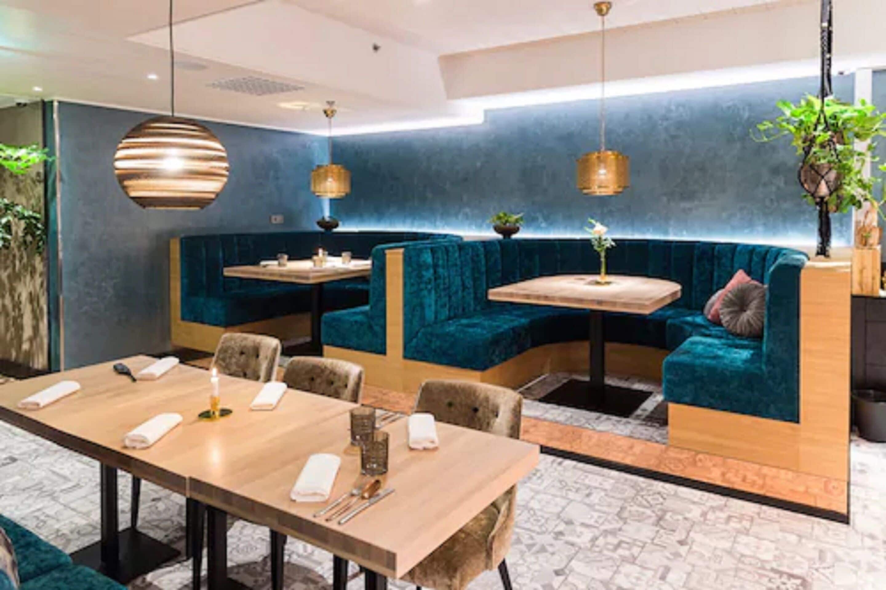 A modern dining area with a long wooden table, cushioned seating, pendant lighting, and a blue accent wall.