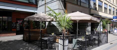 An outdoor cafe with tables and chairs, surrounded by a metal fence, and a building with a 'VERSO' sign in the background.