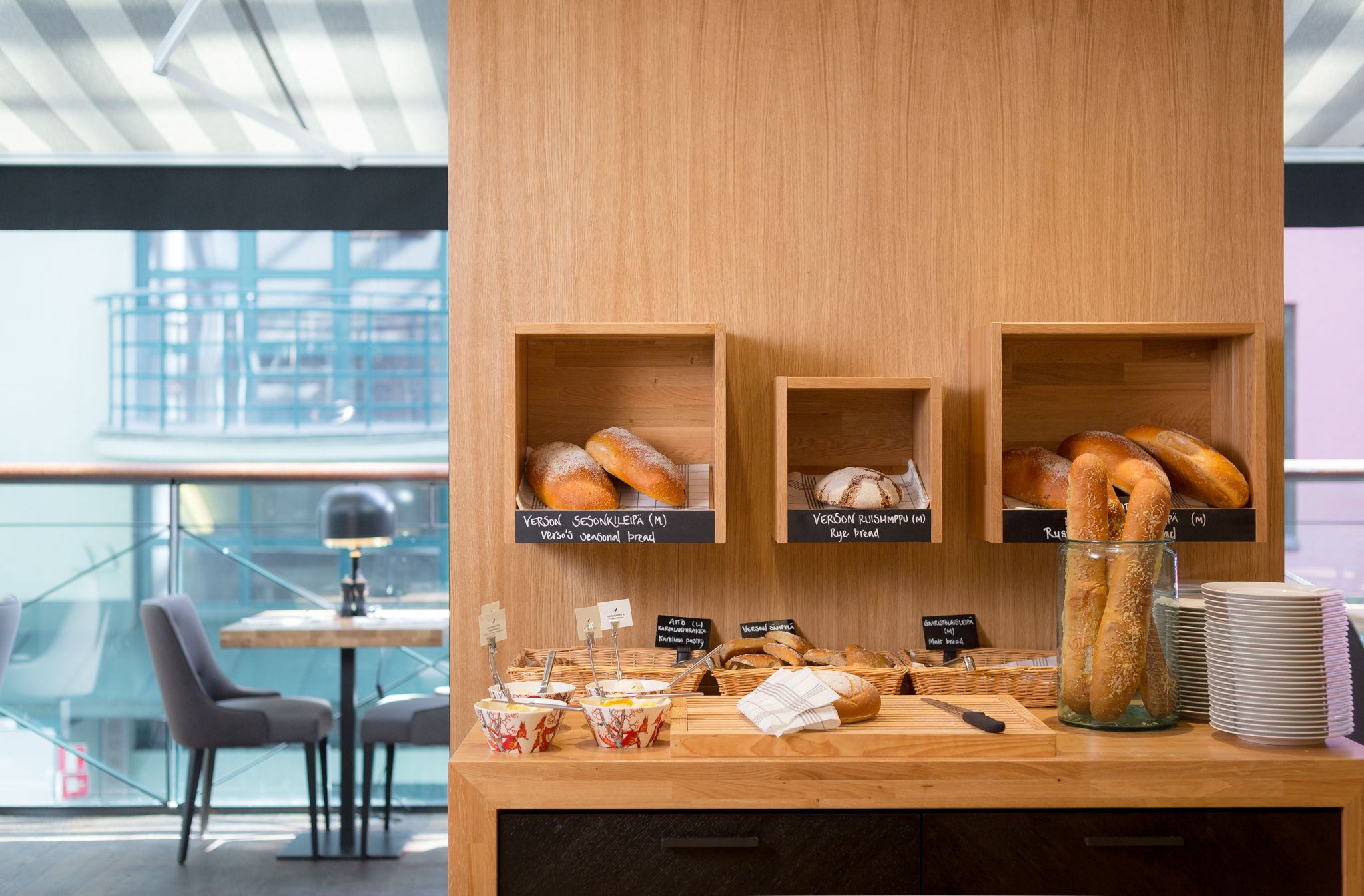 A bakery counter with shelves of assorted breads and pastries, a coffee station, and a view of the cityscape through large windows.