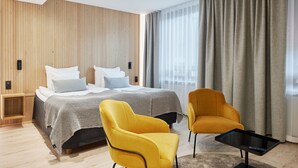 Une chambre d’hôtel moderne avec un mur lambrissé en bois, un grand lit avec une literie blanche, deux fauteuils jaunes et une petite table noire.