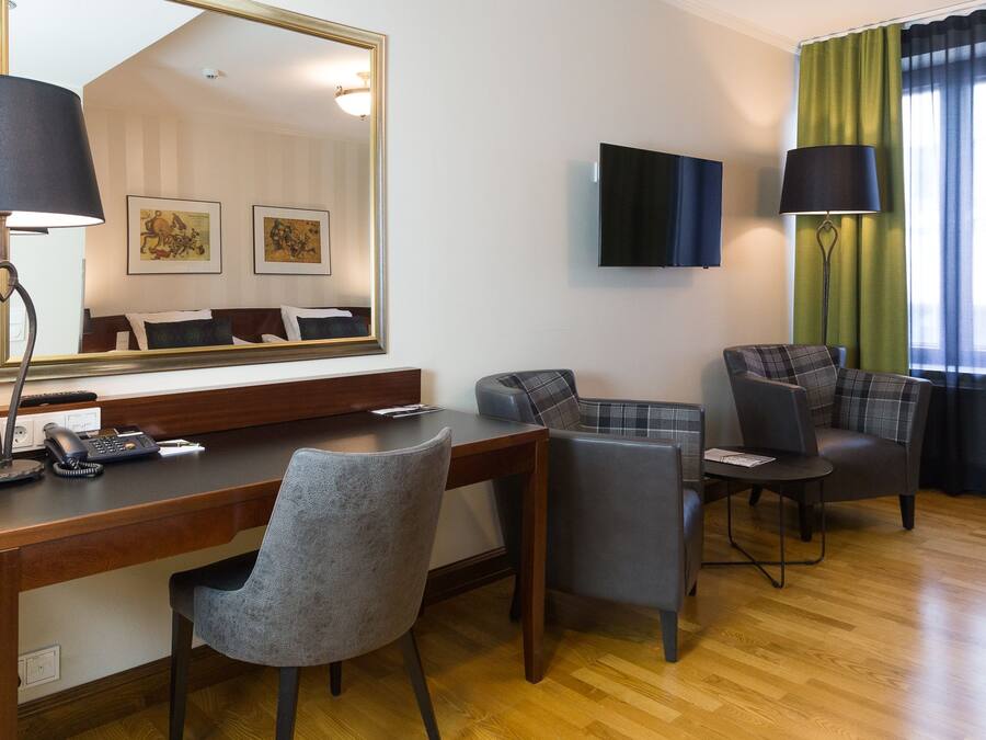 A hotel room with a wooden desk, two grey chairs, a large mirror, a flat-screen TV, and a green curtain.