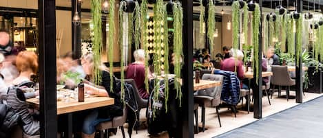 Un comedor moderno con mesas y sillas de madera, plantas colgantes e iluminación colgante.