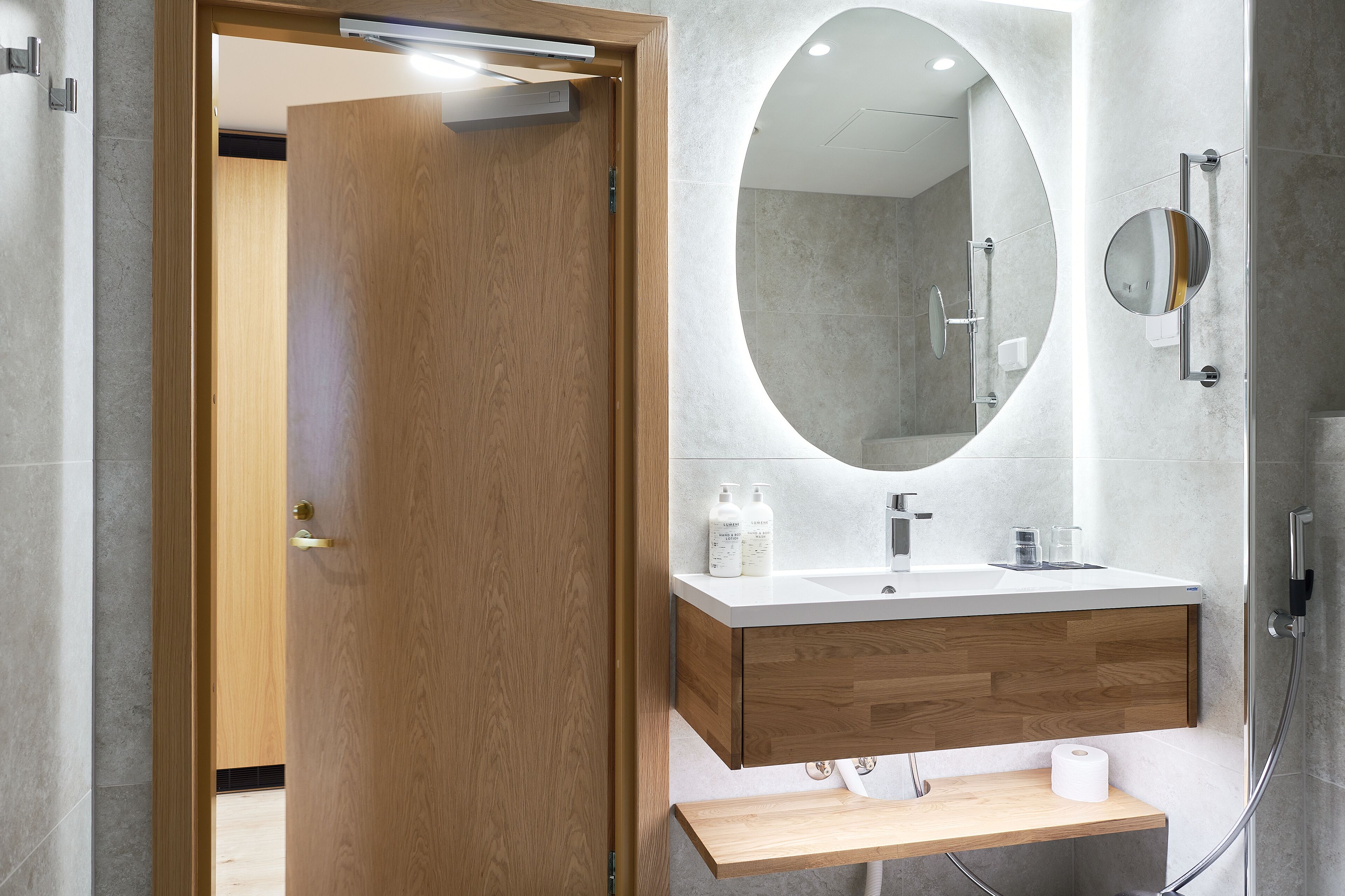 A modern bathroom with a wooden door, a large mirror, a white sink, and a shower area.