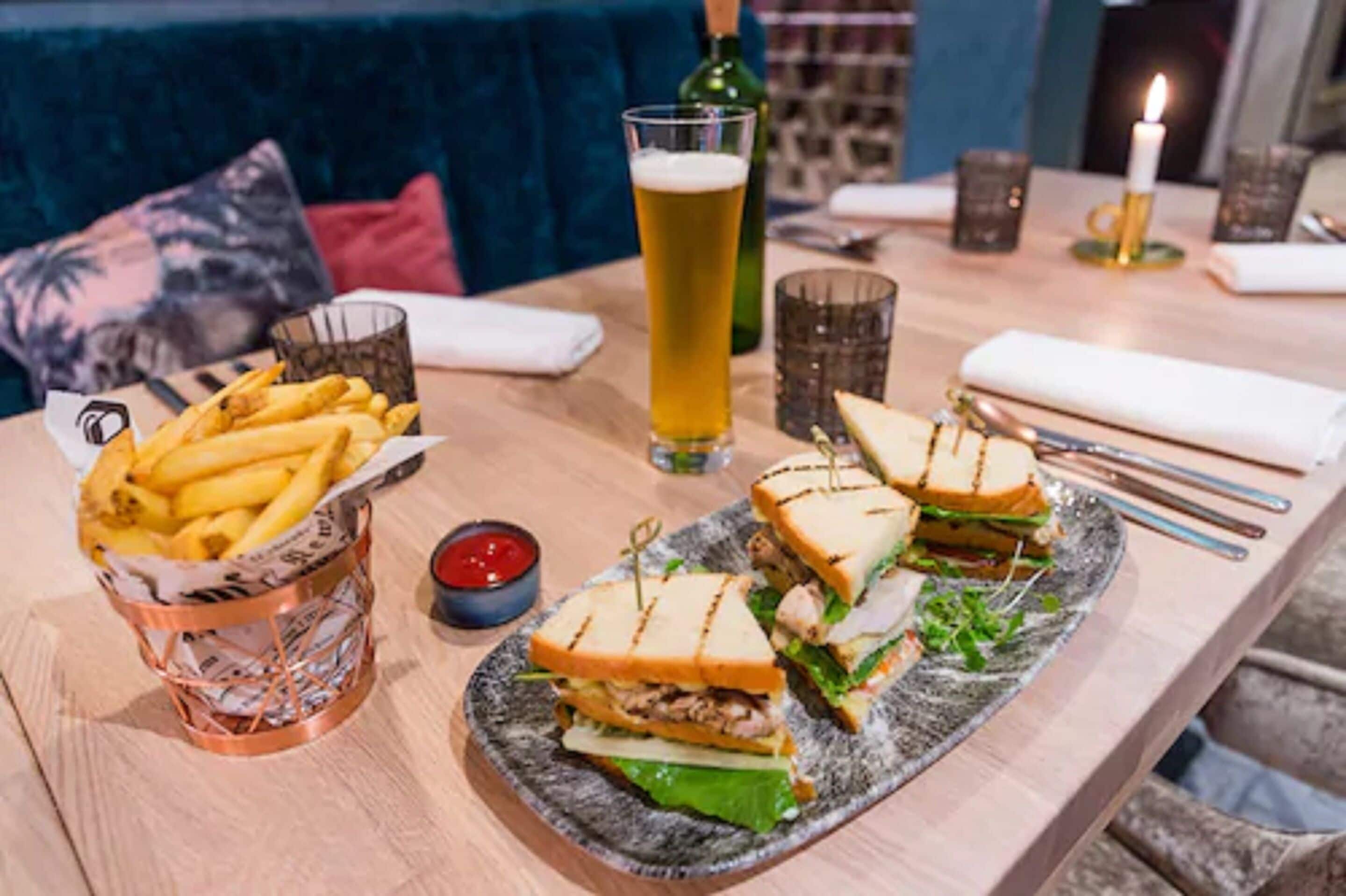 Una mesa con comida que incluye sándwiches, papas fritas y una jarra de cerveza, además de una vela y vasos.