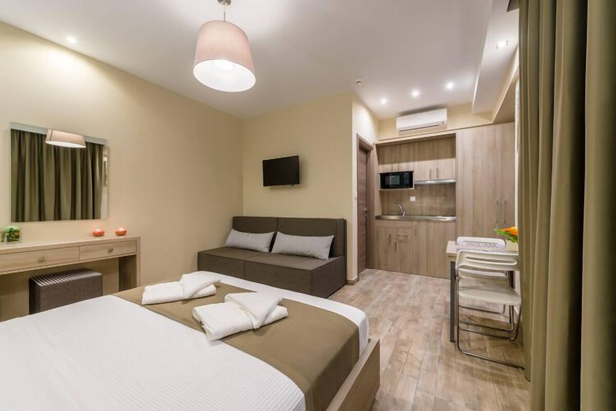 A hotel room with a bed, bedside table, lamp, and a kitchenette area with a microwave and sink.