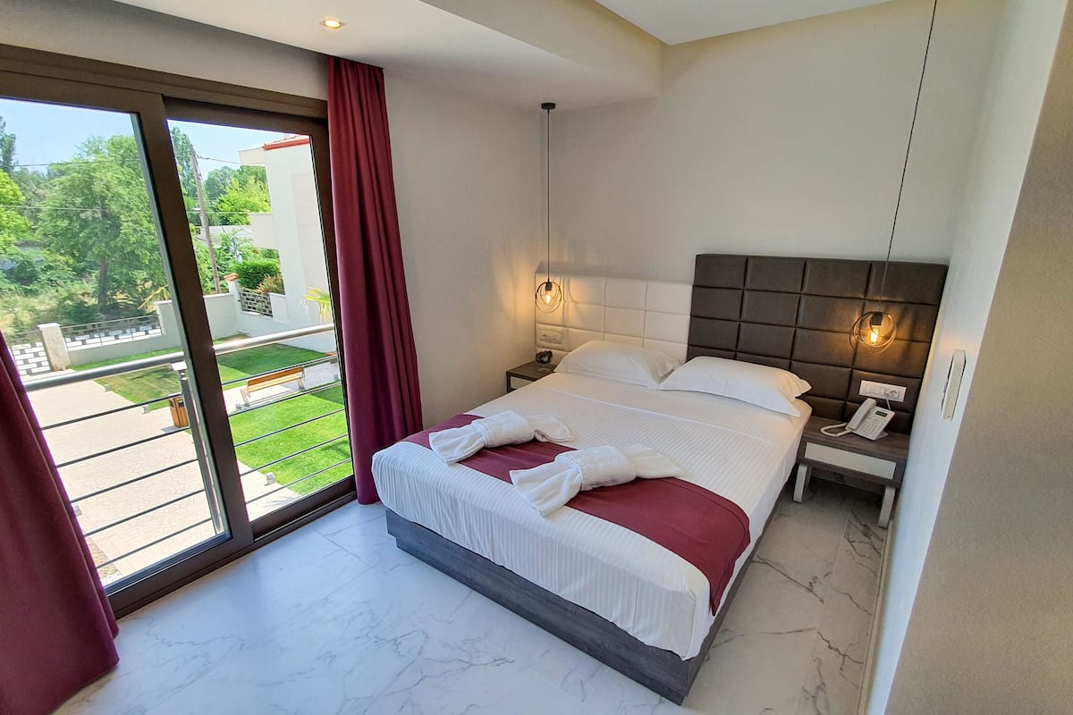 A modern hotel room with a large bed, a bedside table with a phone, and a view of a green garden through a sliding door.