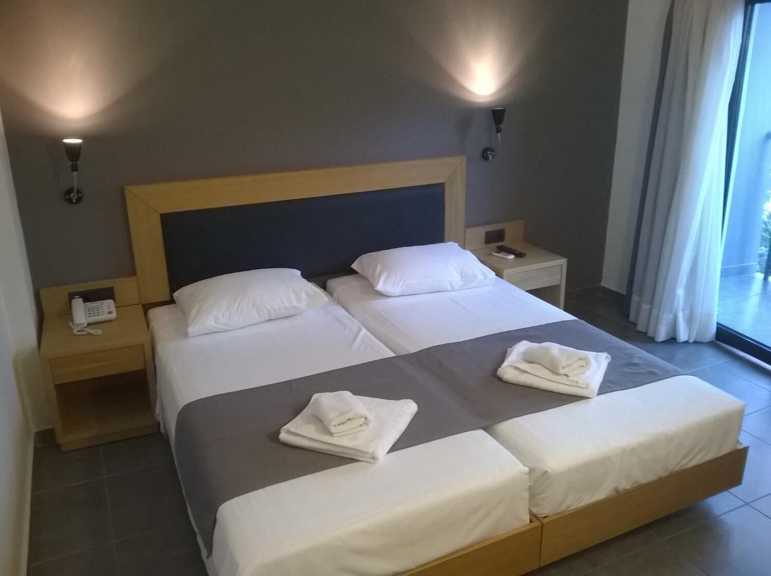Une chambre d’hôtel avec un grand lit, deux tables de chevet, un téléphone sur la table de gauche et une fenêtre avec des rideaux.