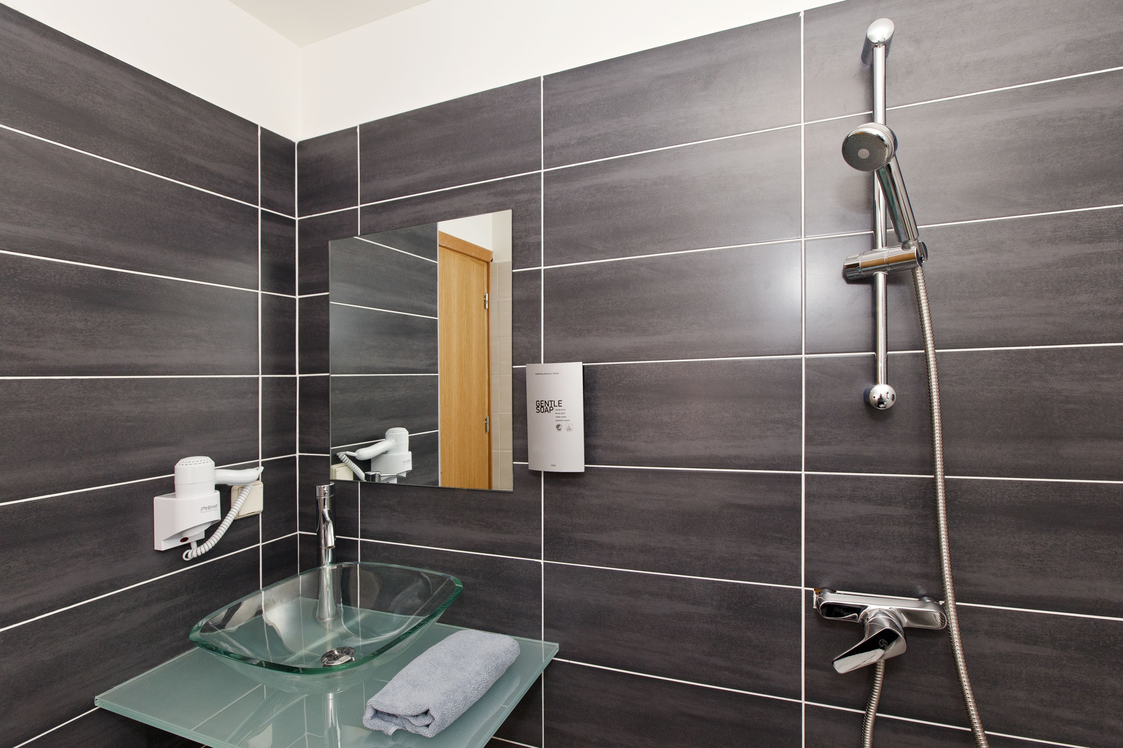 Ein modernes Badezimmer mit dunklen Fliesen, einem Glastischwaschbecken, einer Handbrause und einem Handtuchhalter.