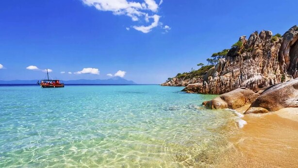 Egy tiszta, homokos strand, a távolban egy hajóval, és sziklás sziklák.