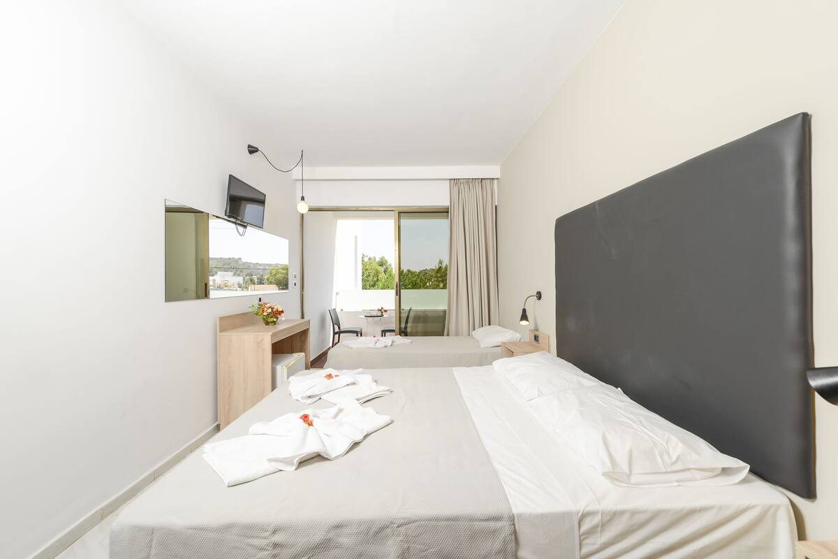 A modern hotel room with a large bed, a desk with a chair, a flat-screen TV mounted on the wall, and a large window with curtains.