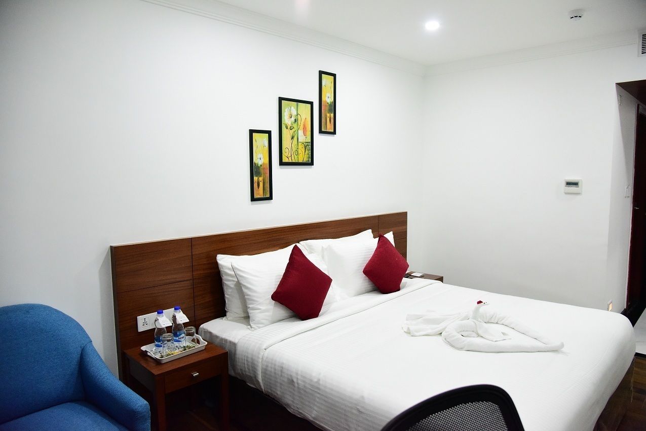 A hotel room with a wooden headboard, white bedding, red pillows, a blue armchair, a bedside table with water bottles, and three framed artworks on the wall.