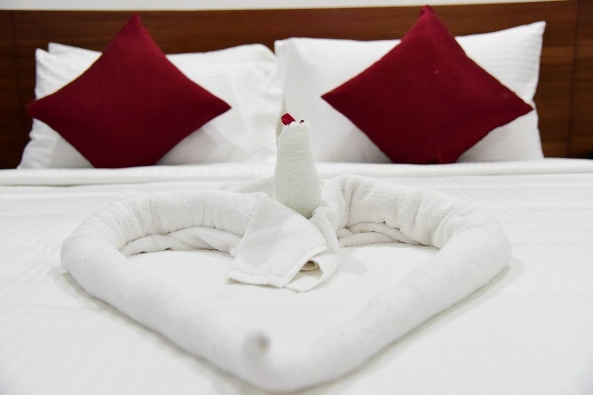 A neatly made bed with a white towel arranged in a swan shape, red pillows, and a wooden headboard.