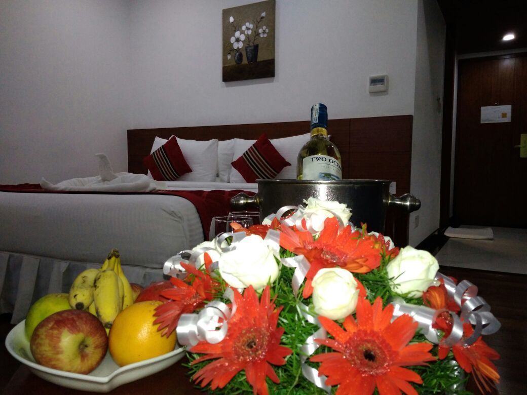 Una habitación de hotel con una cama, una mesita de noche con una botella, un cuenco de fruta y un arreglo floral.