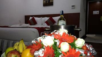 Una habitación de hotel con una cama, una mesita de noche con una botella, un cuenco de fruta y un arreglo floral.