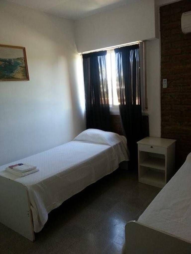 A small, single-bed room with a window, a nightstand, and a wall-mounted air conditioning unit.