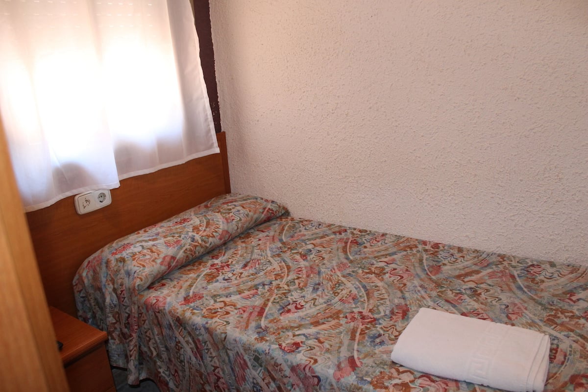 Une petite chambre avec un lit simple, une couvre-lit à motifs, une tête de lit en bois et une serviette blanche sur le lit.