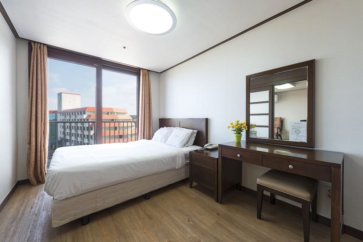 Habitación de hotel con cama, escritorio, silla, espejo y ventana con cortinas.