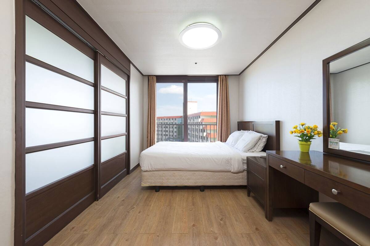 Habitación de hotel con cama, cómoda, espejo, planta en maceta y vistas a edificios a través de una ventana.