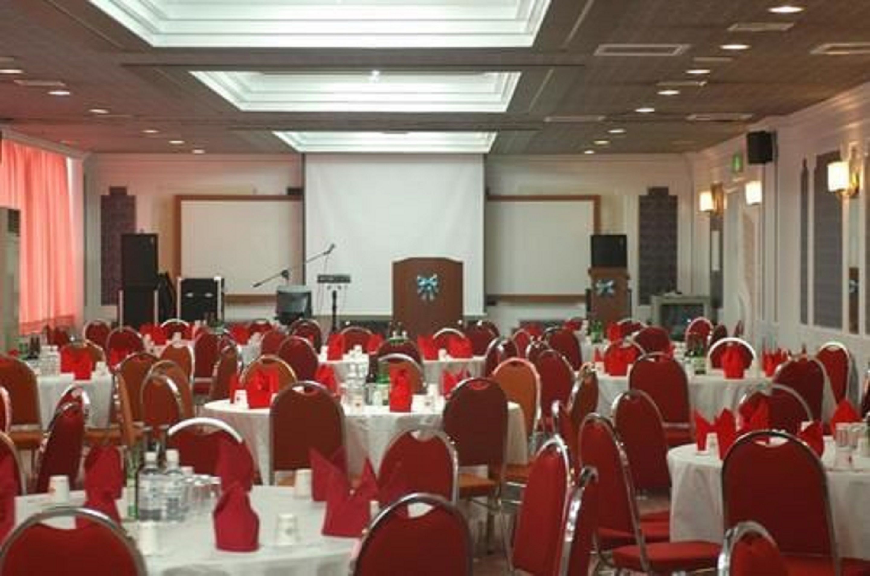 Un salón de banquetes con mesas redondas, sillas rojas y un escenario con un atril.