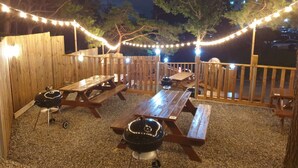 Een terras buiten met houten banken en picknicktafels, lichtjes en een barbecue.