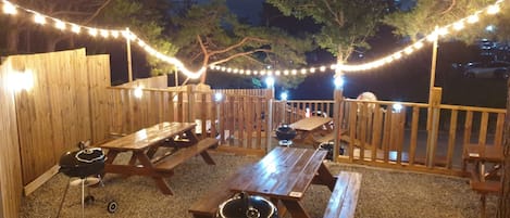 An outdoor patio area with wooden benches and picnic tables, string lights, and a barbecue grill.