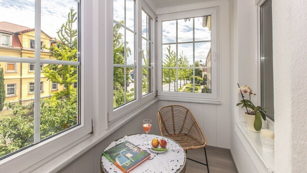 Una camera d'angolo con ampie finestre, una sedia in vimini, un tavolino con un libro e della frutta, e un'orchidea in vaso.
